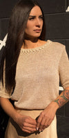 Woman wearing a gold knit top with pearl details, standing outdoors in sunlight