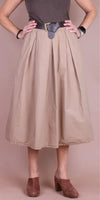 Woman wearing a beige midi skirt with pleats, brown belt, and brown heeled mules.