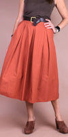 Woman wearing a rust-colored midi skirt with pleats, black belt, and brown heeled shoes