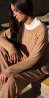 Woman in tan long sleeve top and pants with white ribbed neckline, sitting outdoors in sunlight