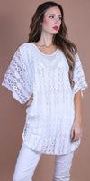 Woman wearing a white lace tunic top with batwing sleeves and white pants, neutral background