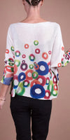 Woman wearing white knit sweater with colorful circular pattern, black pants, neutral background