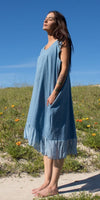 Woman in blue sleeveless linen dress with lace trim, standing barefoot outdoors on a sunny day