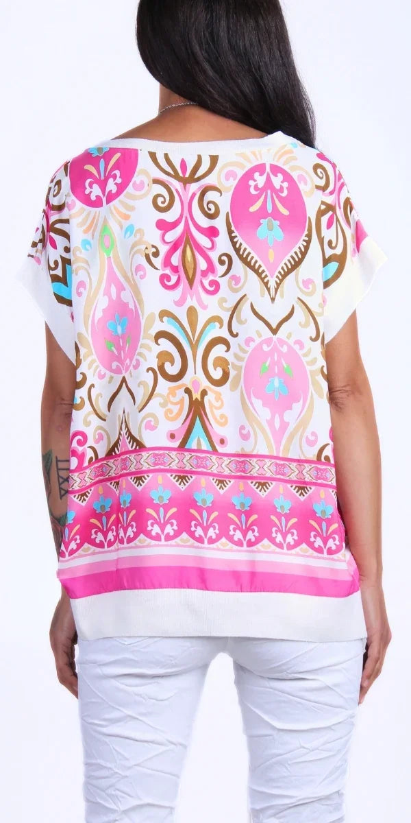 Back view of woman wearing a white top with pink, gold, and blue baroque print and white pants